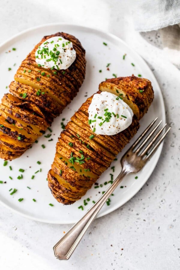 2 hasselback potatoes with sour cream and chives on top on a plate with a fork