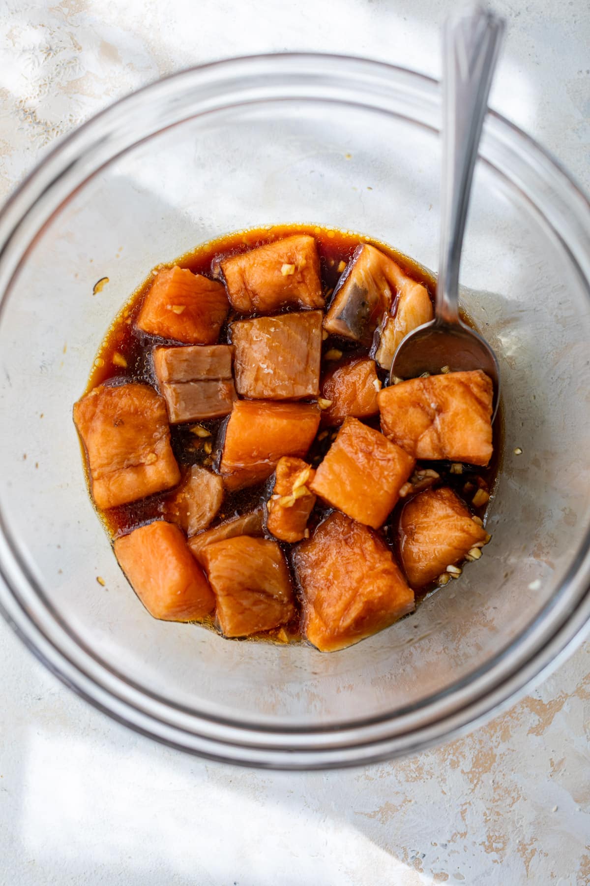 using a spoon to toss salmon bites in a soy sauce marinade in a glass bowl.