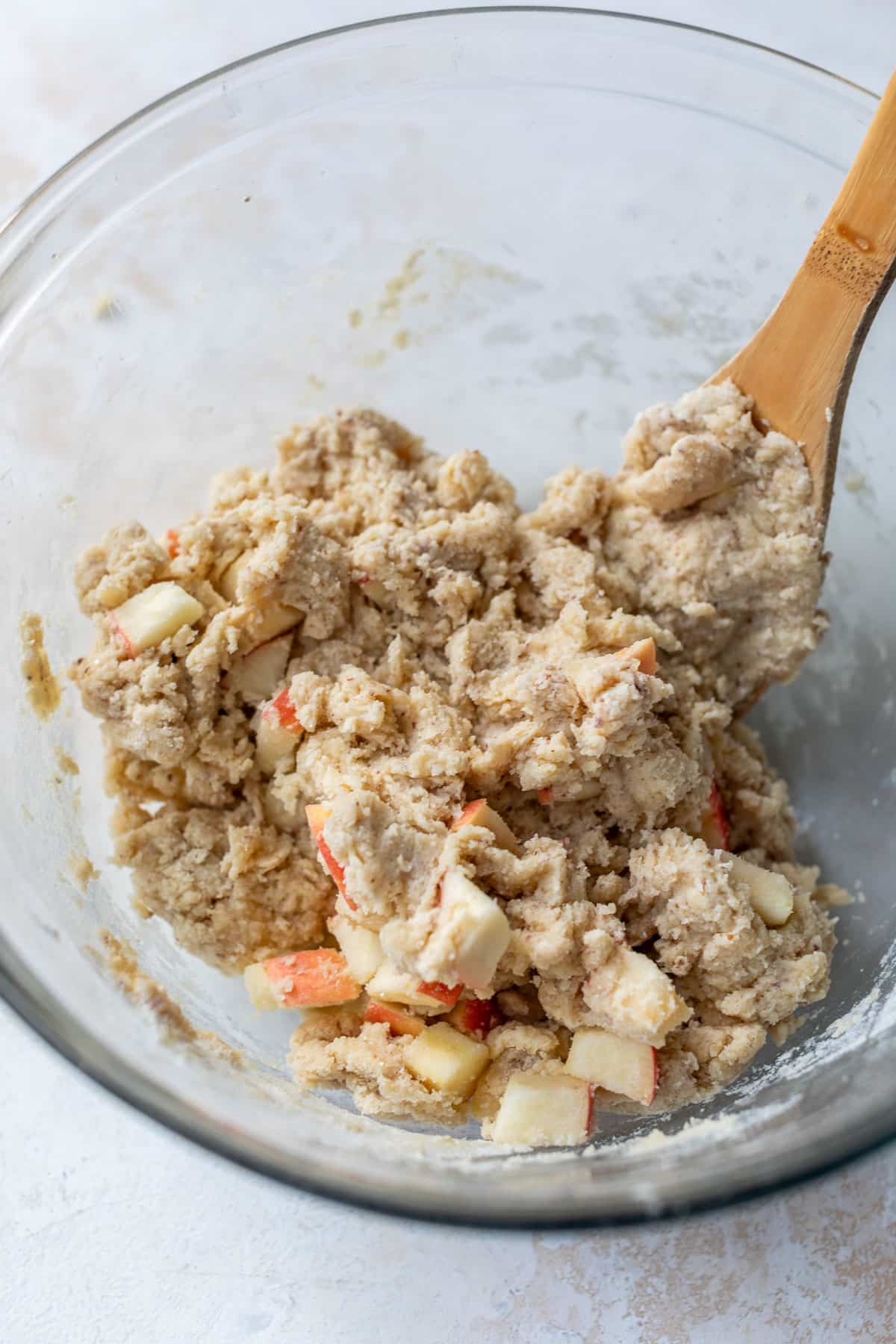 using a wooden spoon to fold chopped apples into apple cranberry scone dough in a glass bowl.