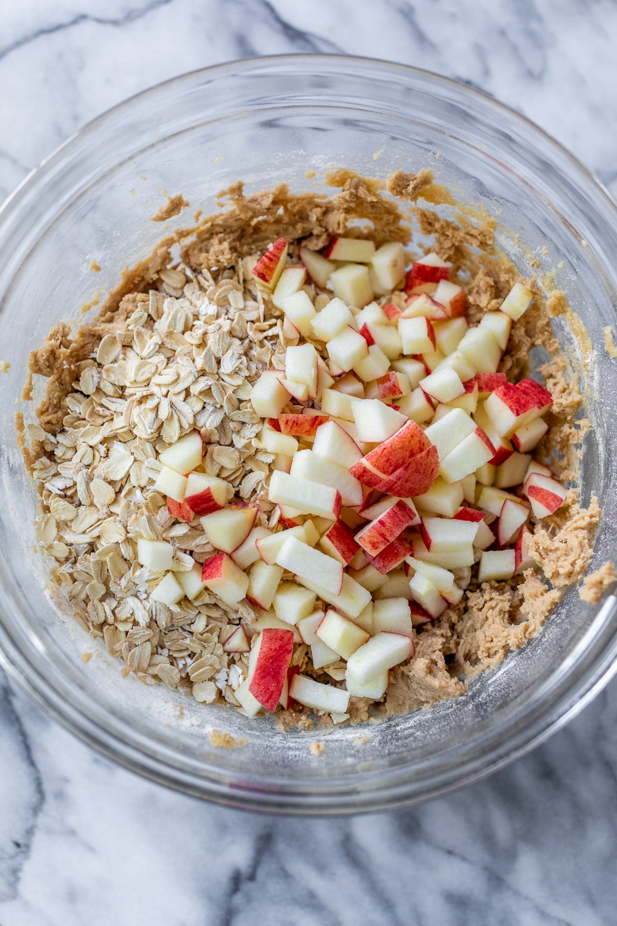 apple chunks and oats over top of apple oatmeal cookie dough in a glass bowl.