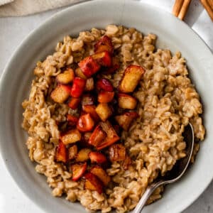 A gray colored bowl beside a cinnamon stick and a linen. Inside the bowl is cooked oatmeal topped with diced cooked apple.