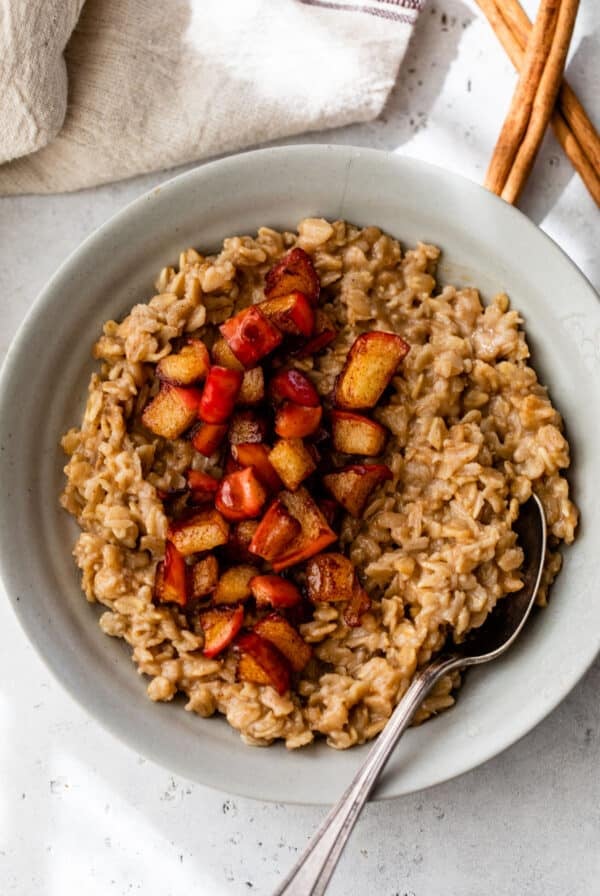 A gray colored bowl beside a cinnamon stick and a linen. Inside the bowl is cooked oatmeal topped with diced cooked apple.