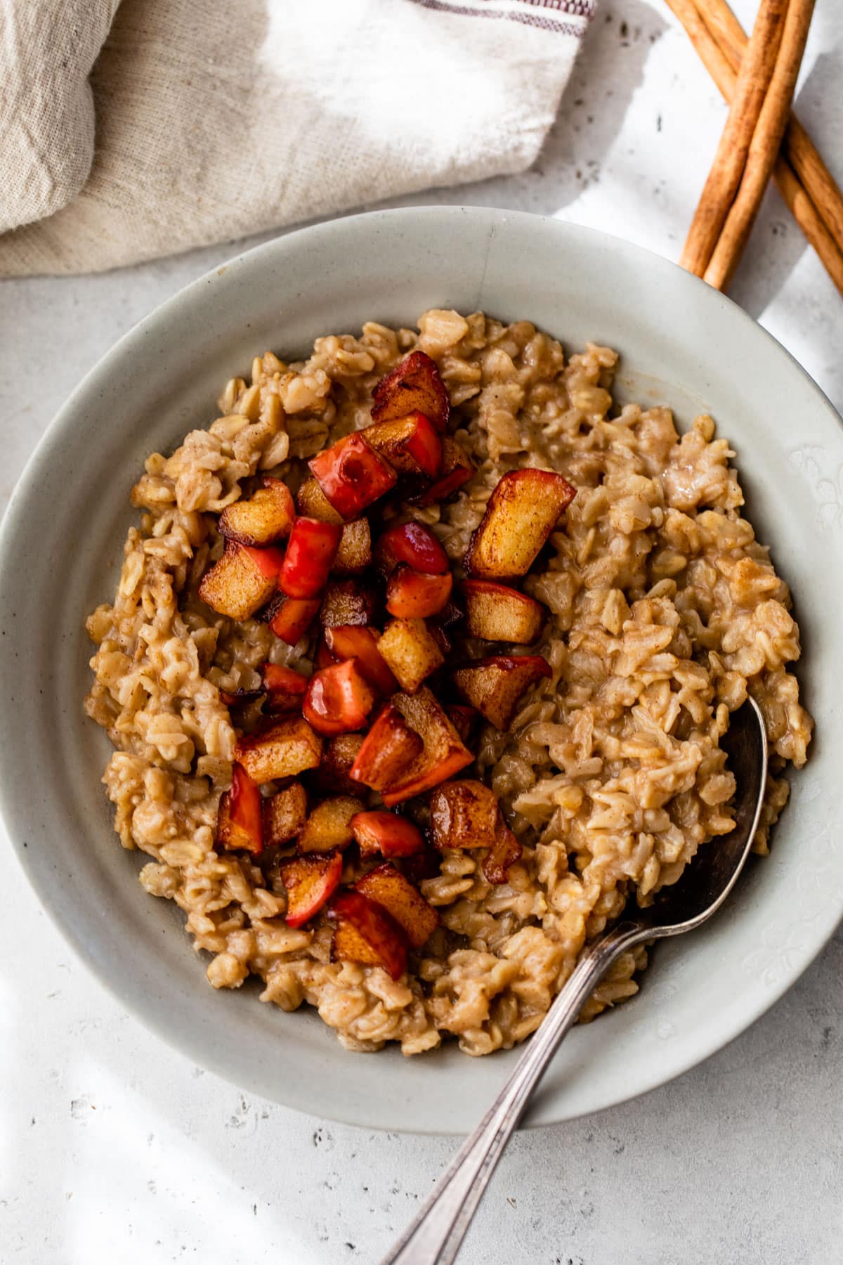 A gray colored bowl beside a cinnamon stick and a linen. Inside the bowl is cooked oatmeal topped with diced cooked apple.