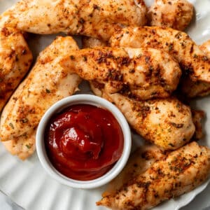 a pile of baked chicken tenders on a white plate next to a small bowl of ketchup.