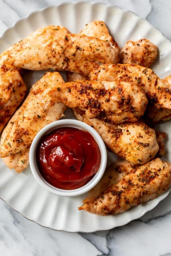 a pile of baked chicken tenders on a white plate next to a small bowl of ketchup.