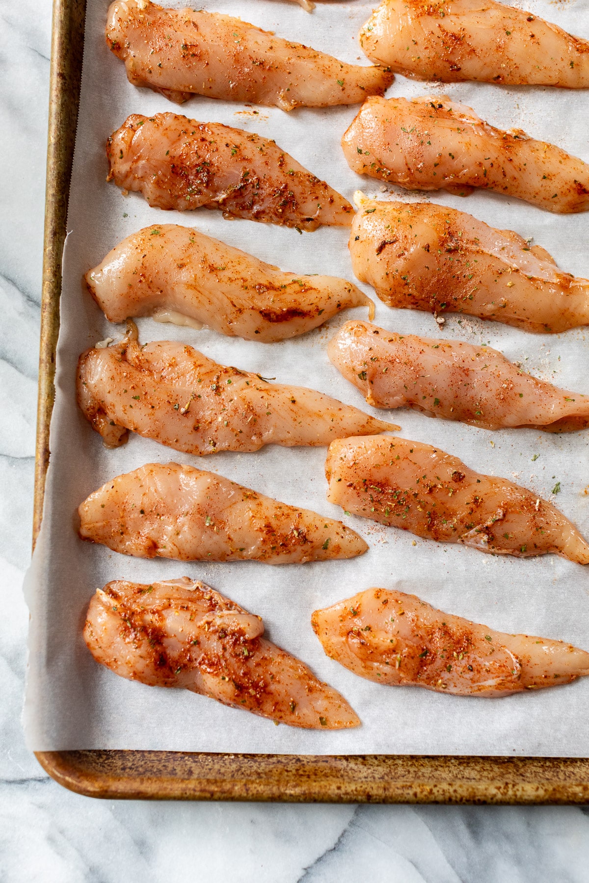 raw seasoned chicken tenders in rows on a baking sheet.