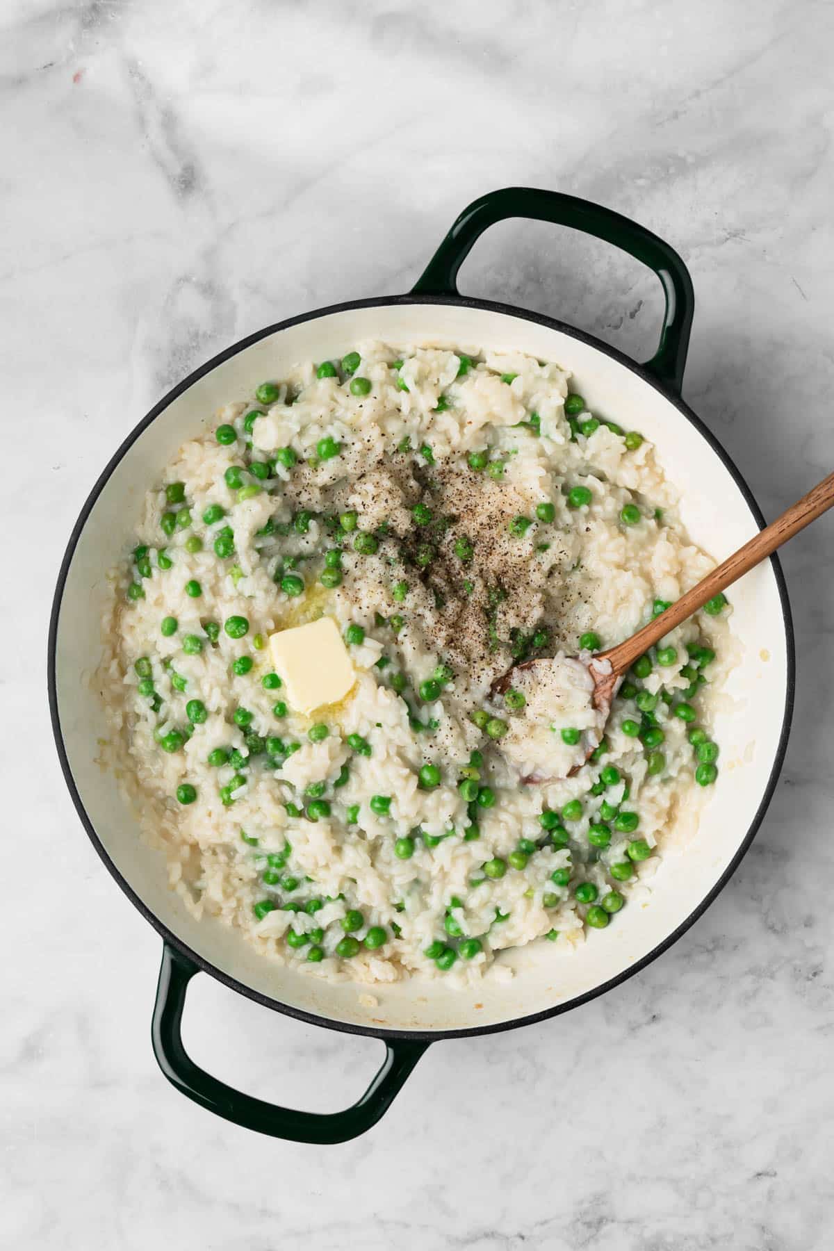overhead view of a wooden spoon stirring butter, salt, and pepper into a pot of baked risotto.