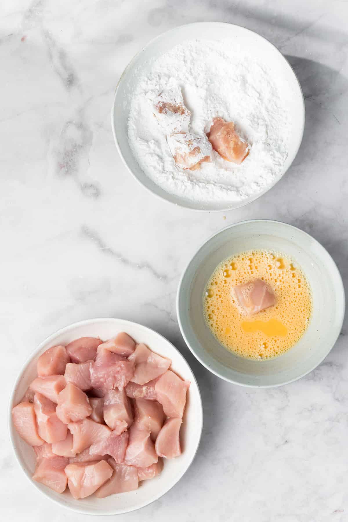 overhead view of raw chicken pieces, whisked eggs, and flour in 3 individual white bowls.