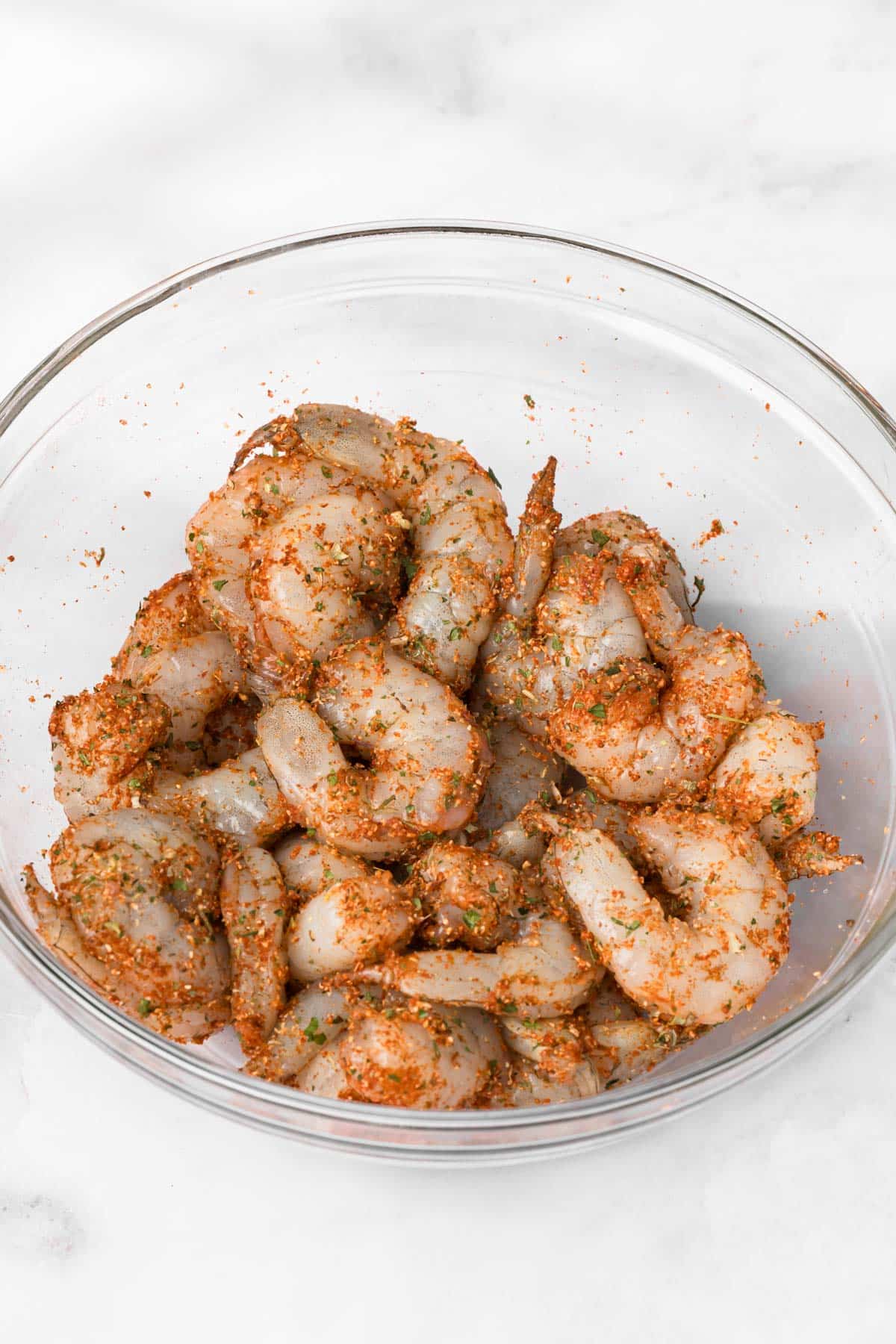 overhead view of raw shrimp covered in seasonings in a glass bowl.