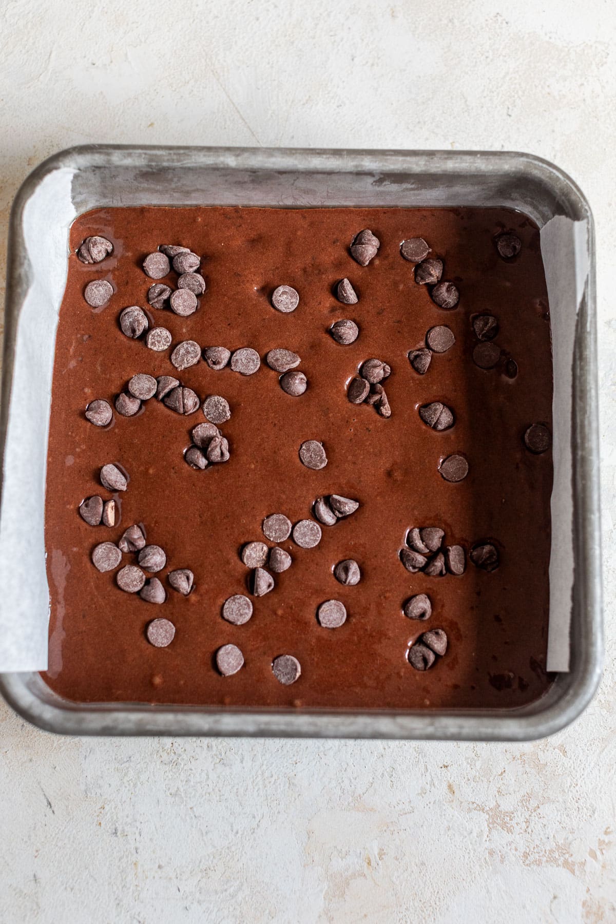 overhead view of black bean brownie batter topped with chocolate chips in a lined baking dish.