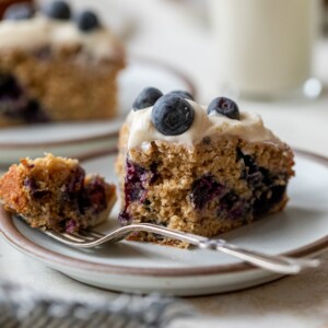 side view of a slice of blueberry cake on a plate with a fork and with a bite taken out