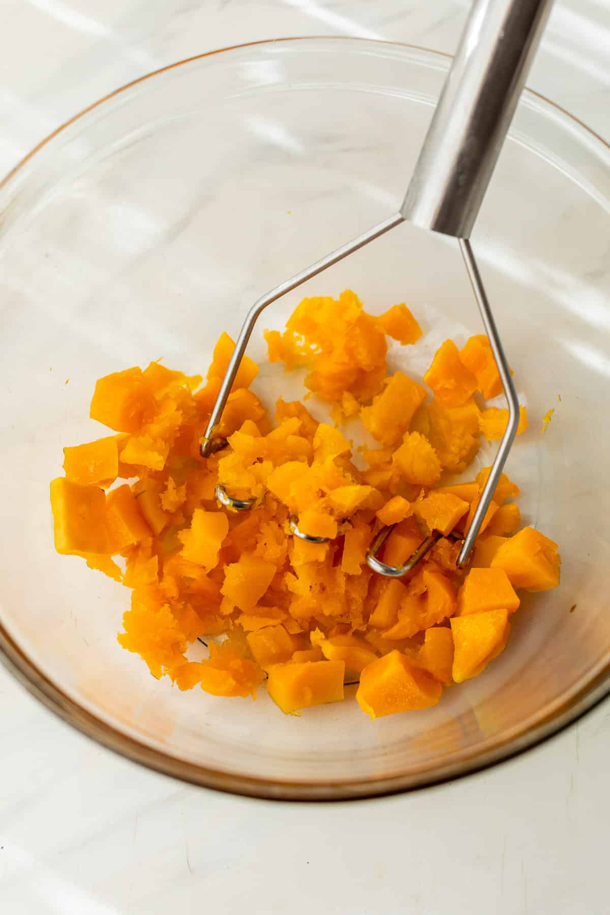 mashing butternut squash cubes in a glass bowl with a potato masher.
