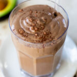 chocolate smoothie in a glass with a straw beside an avocado
