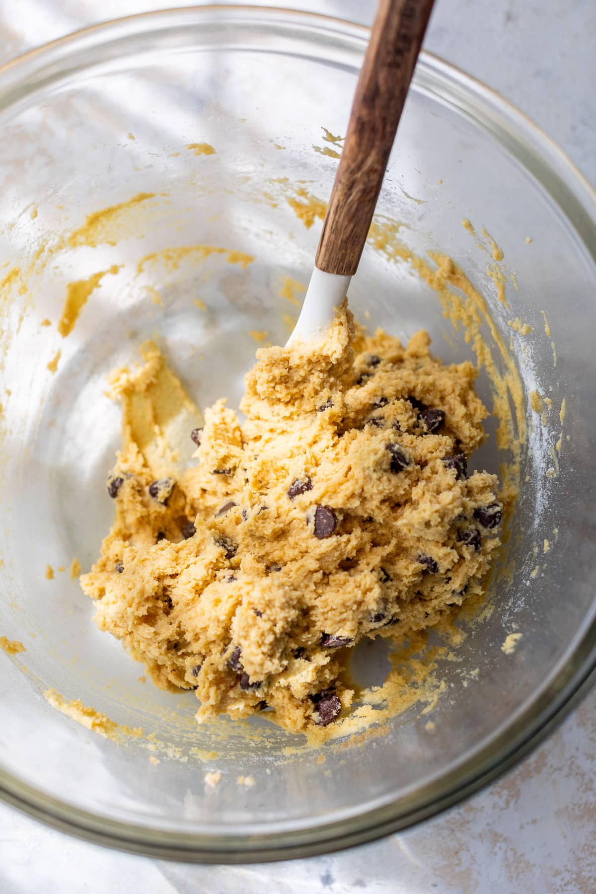 using a spatula to fold chocolate chips into coconut flour cookie dough in a large glass bowl.