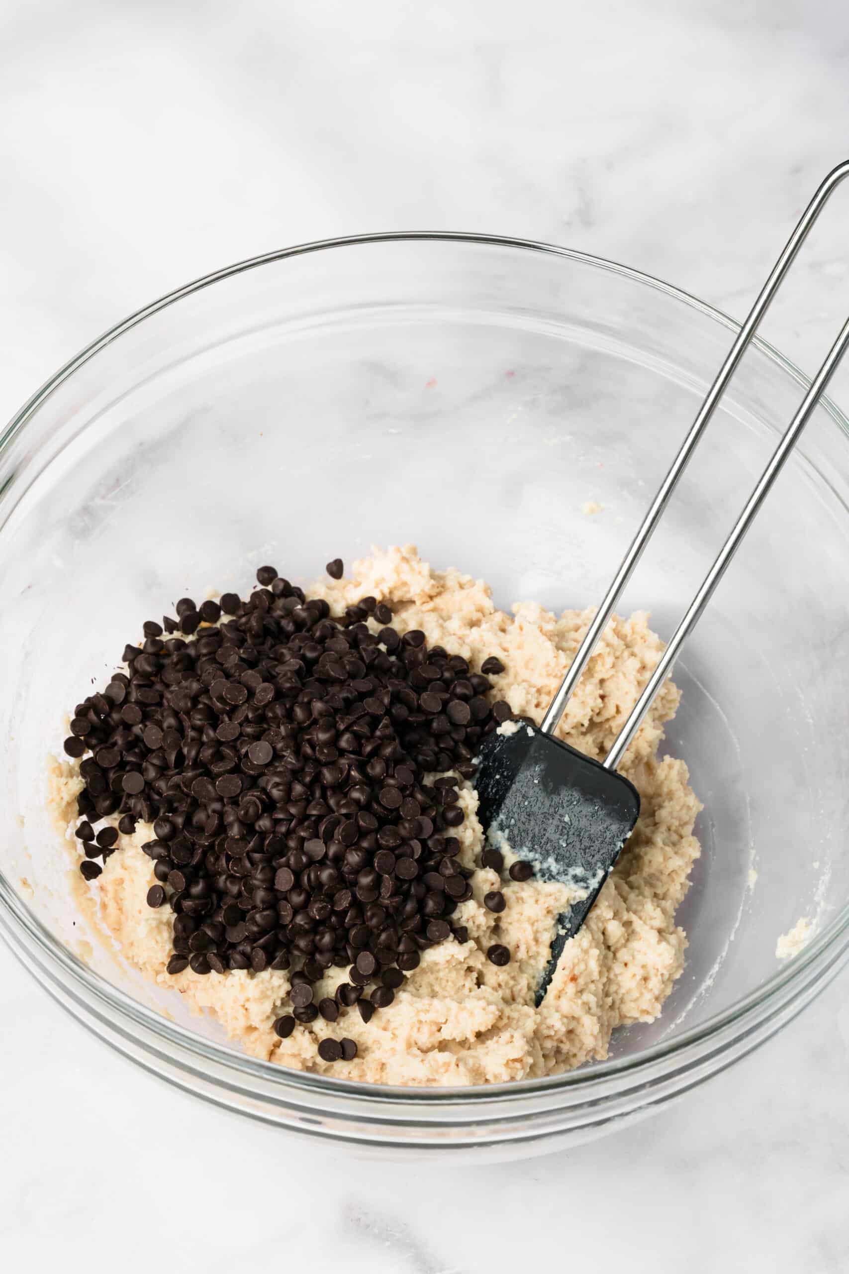 side view of a black spatula mixing chocolate chips into cottage cheese cookie dough in a glass bowl.