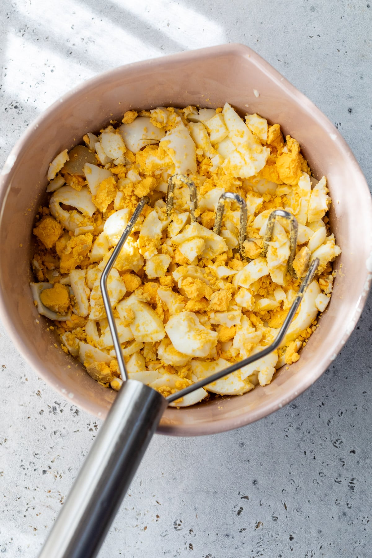 mashing hard boiled eggs with a potato masher in a large pink bowl.