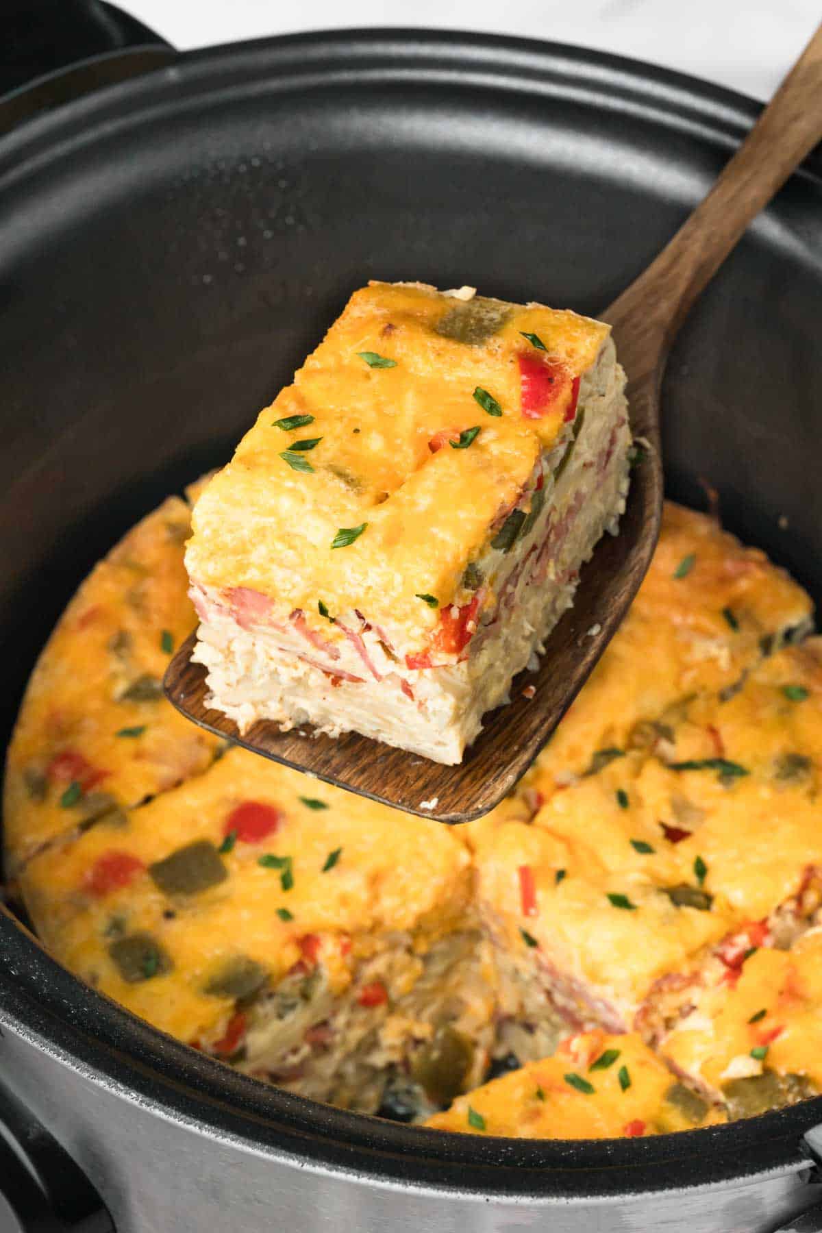side view of a wooden spoon lifting a slice of a cooked crockpot breakfast casserole from a crockpot.