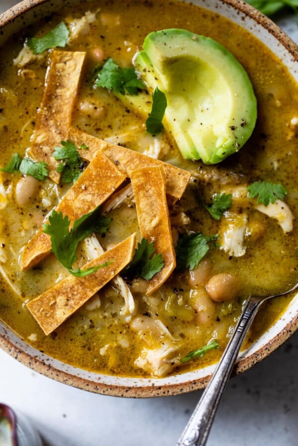 close-up of green chicken chili in a bowl with a spoon and with sliced avocado and tortilla strips on top