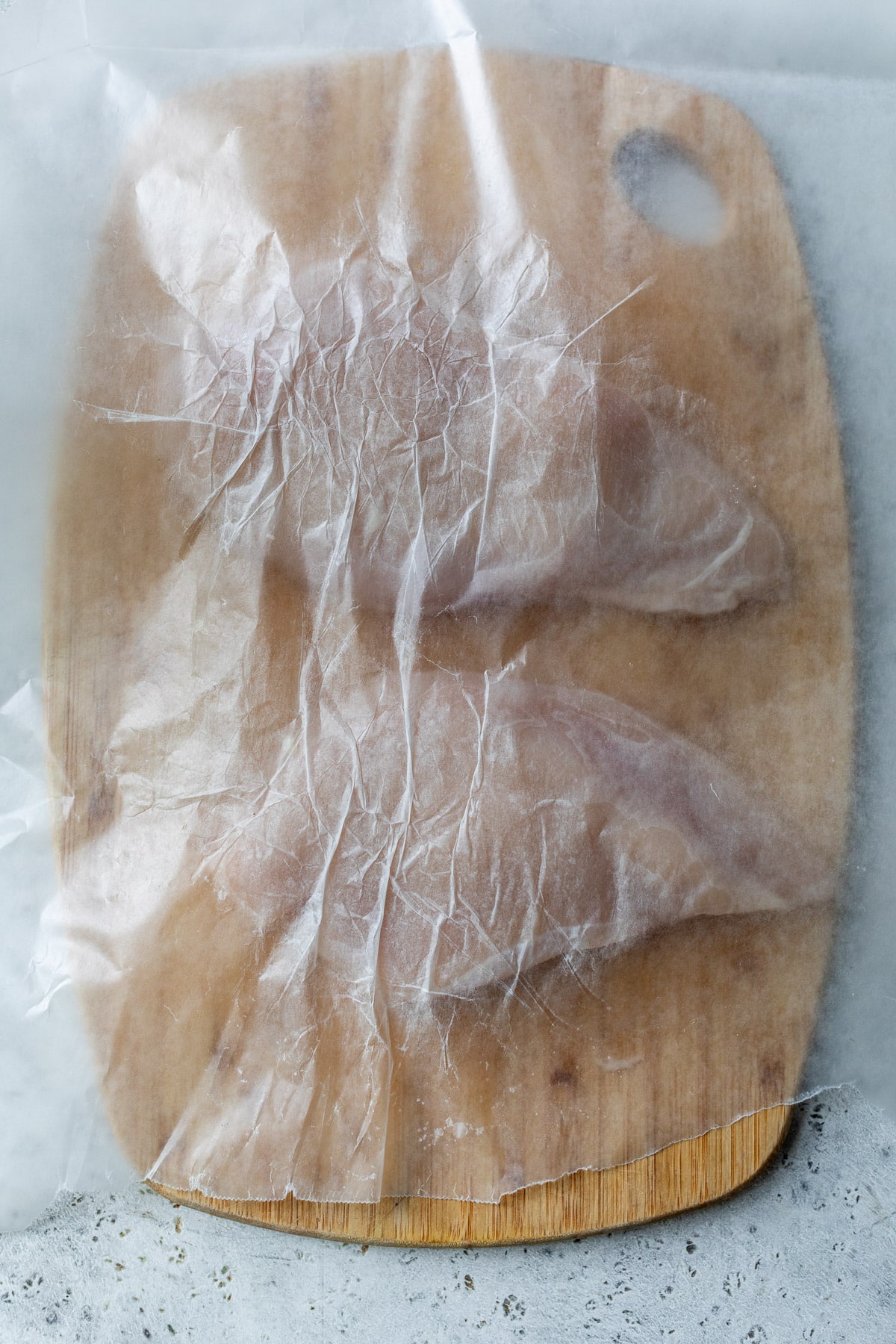 two chicken breasts on a wooden cutting board with a sheet of wax paper on top.