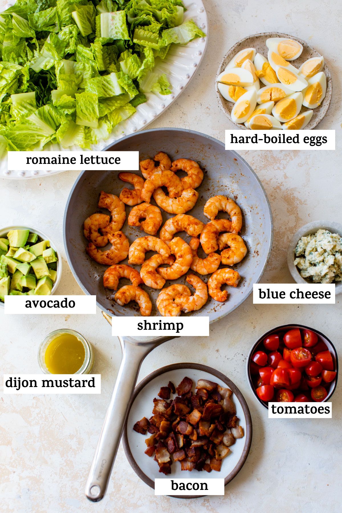 all ingredients on a table; romaine lettuce, hard-boiled eggs, avocado, shrimp, blue cheese, dijon mustard, bacon, and tomatoes