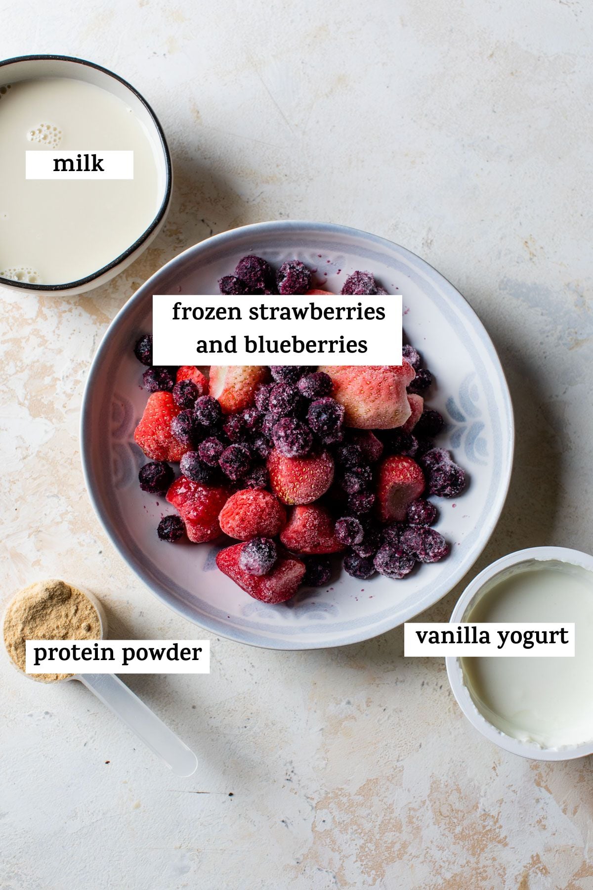 all ingredients on a table and listed out: milk, frozen strawberries and blueberries, protein powder, and vanilla yogurt