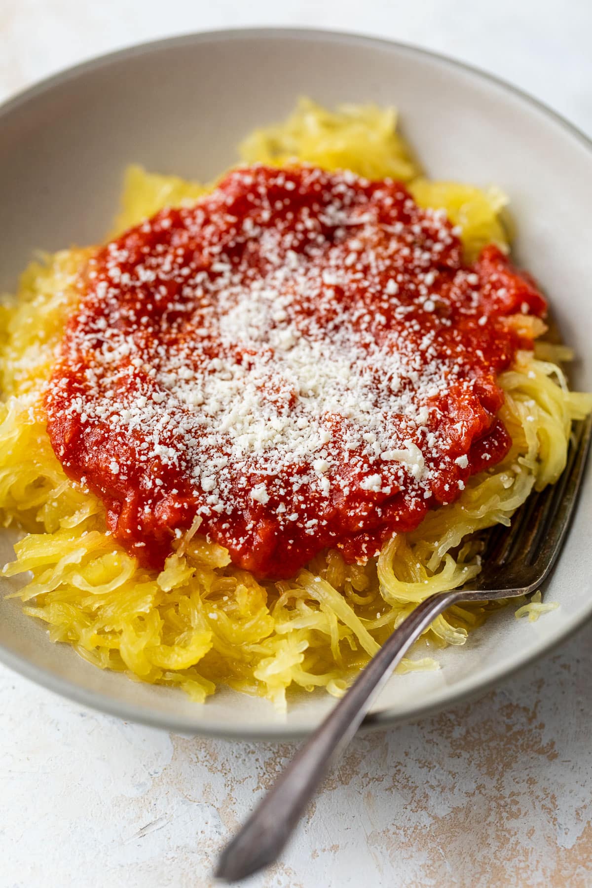 side view of cooked spaghetti squash stands topped with tomato sauce and parmesan in a bowl with a fork.