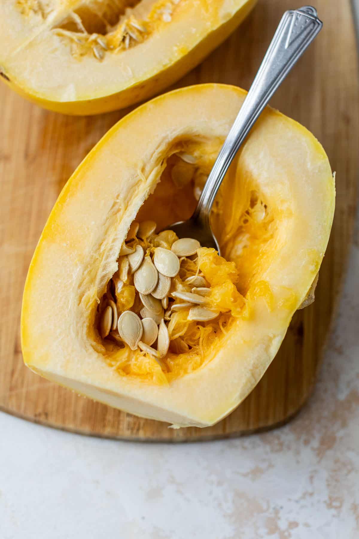 overhead view of a spoon scooping out the seeds from a sliced spaghetti squash on a bamboo cutting board.