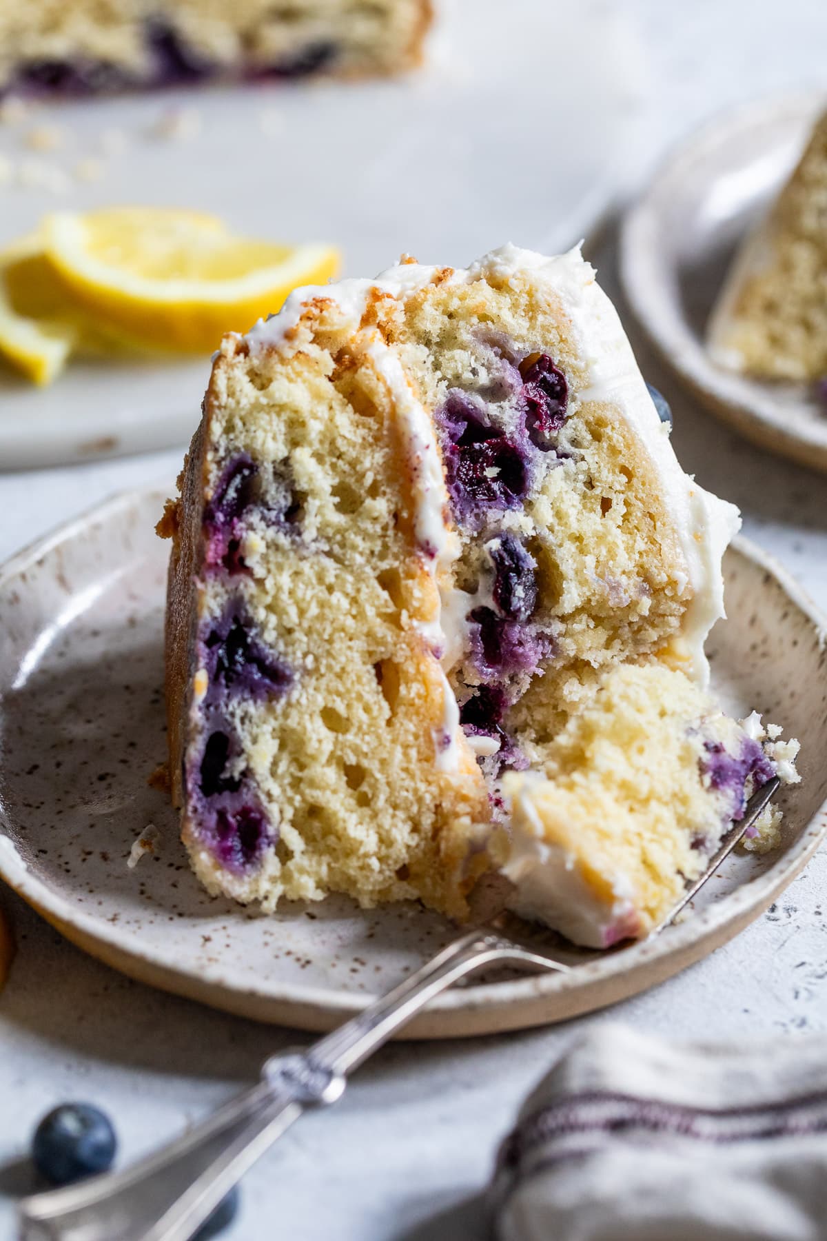 a fork taking a bite out of a slice of lemon blueberry cake on a small plate.