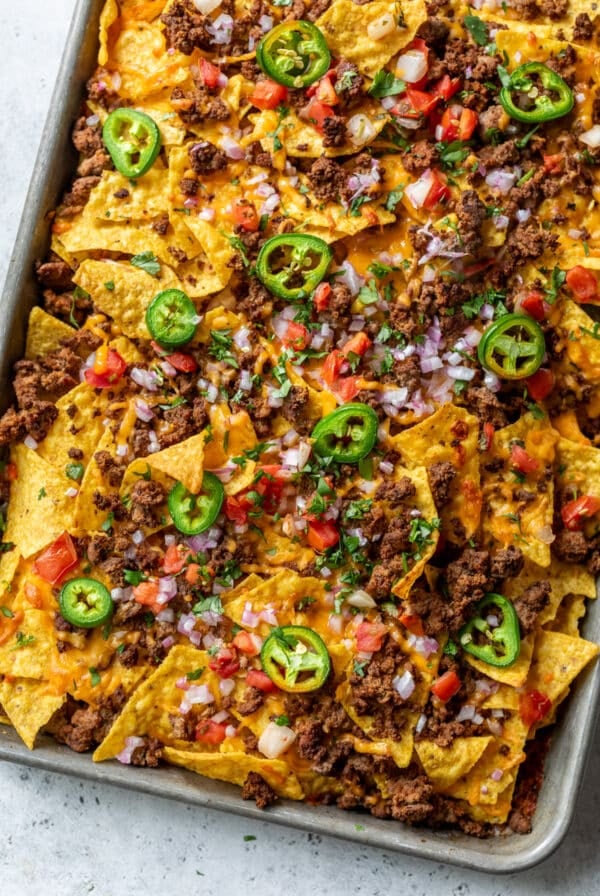 corn tortilla chips on a baking sheet topped with cooked ground beef, diced red onion, diced tomato, cheese, and sliced jalapeño