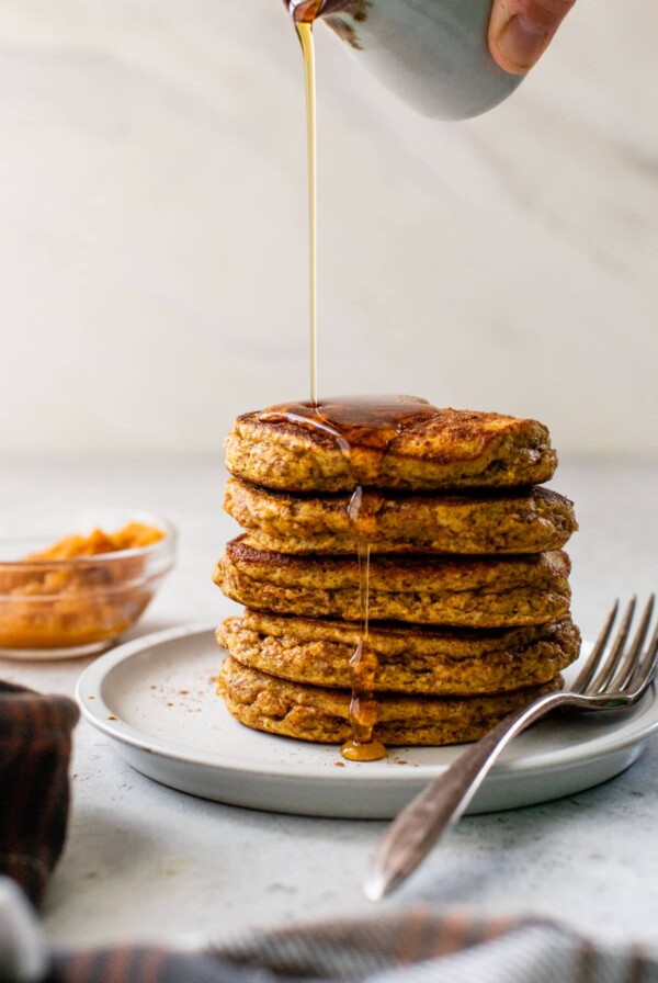 a stack of paleo pumpkin pancakes on a plate with a fork and syrup being poured over the top
