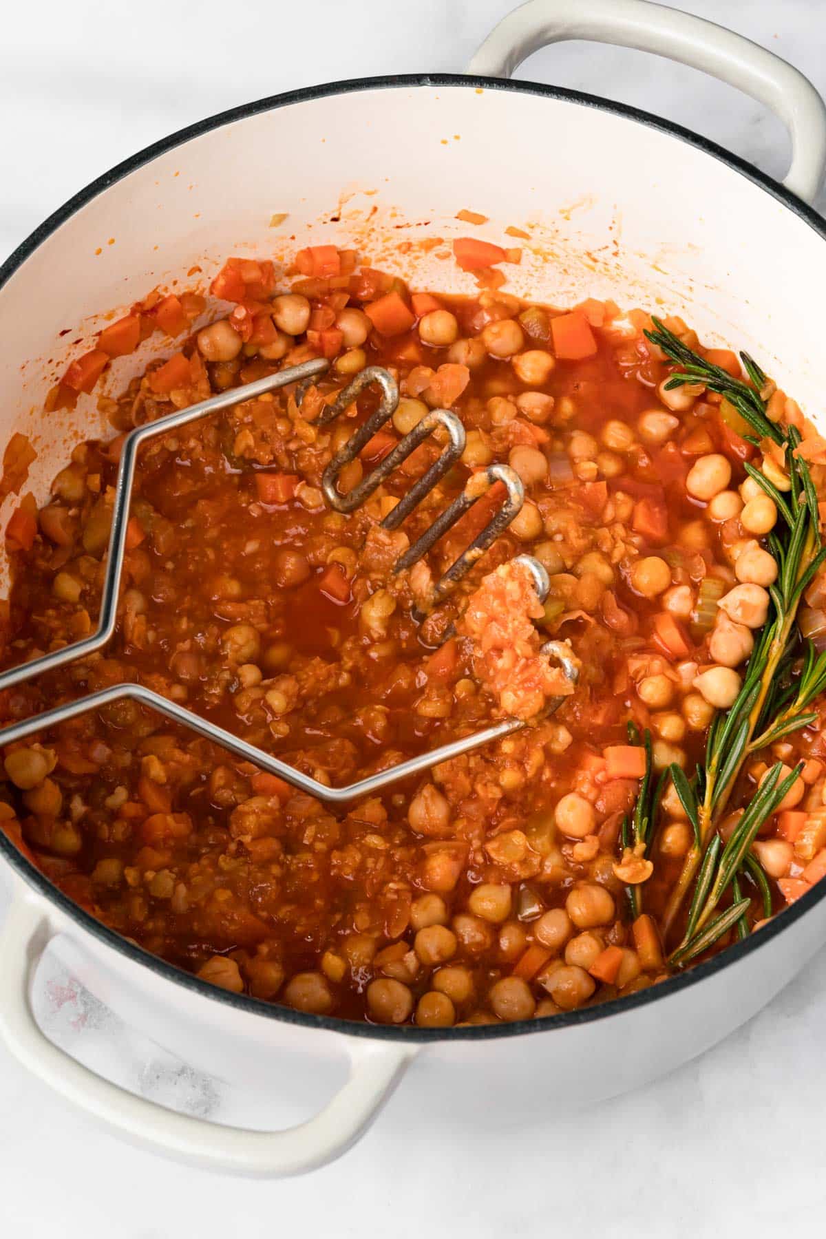 overhead view of a potato masher mashing chickpeas in a pot of pasta e ceci.