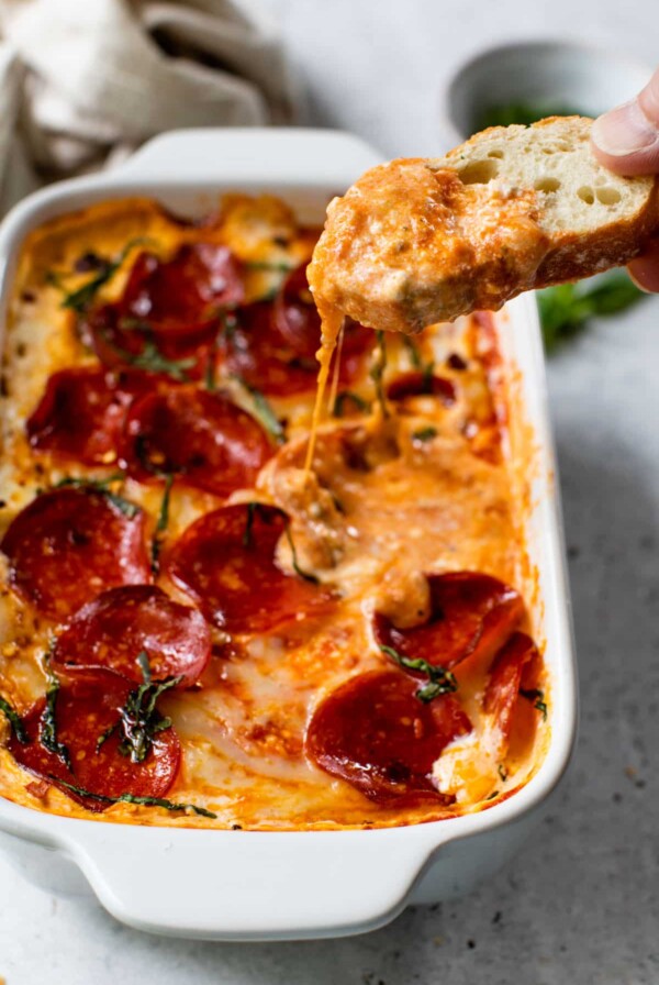 pizza dip in a baking dish with a slice of bread dipped into the dip and with chopped fresh basil on top