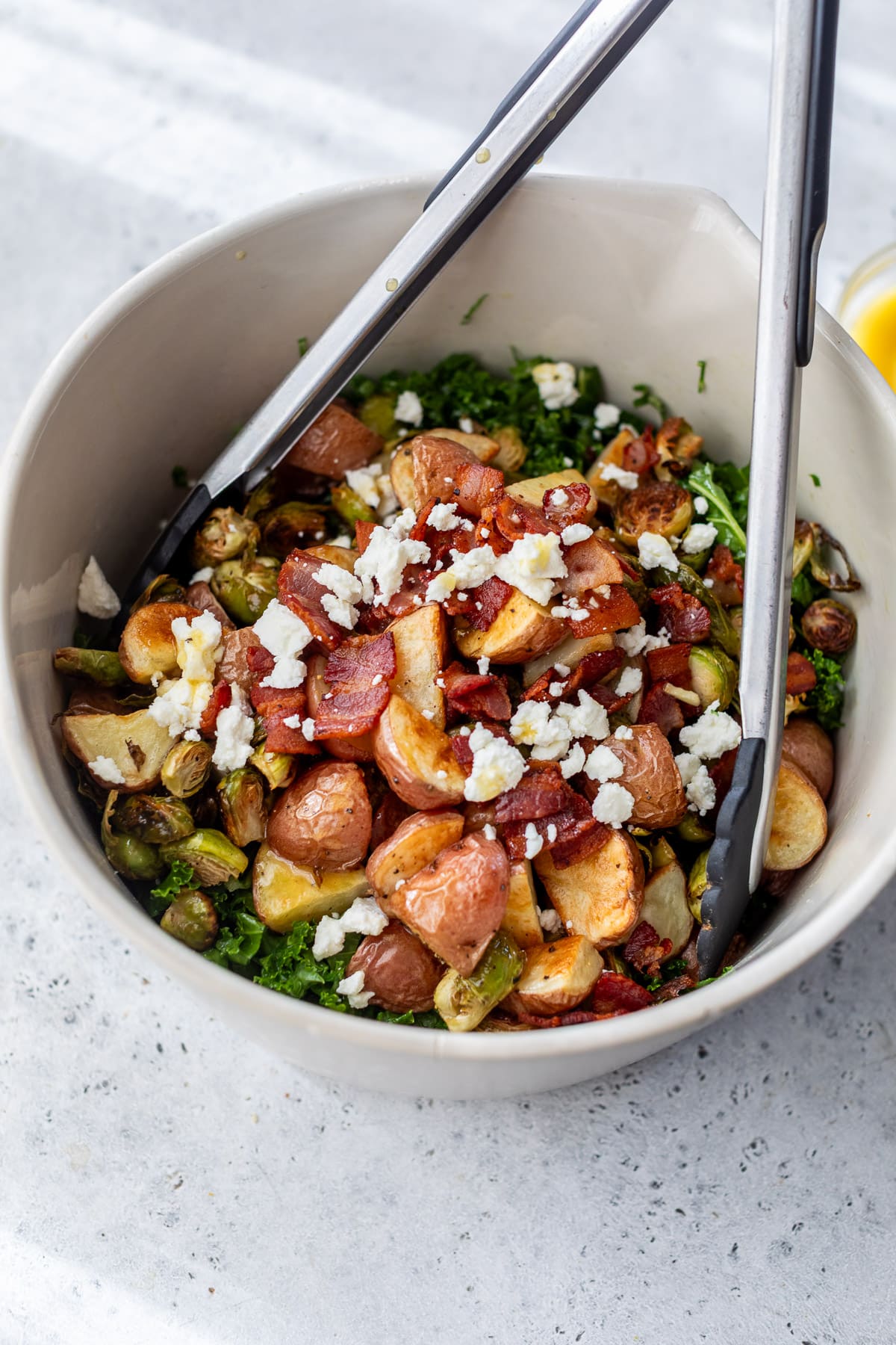 using tongs to mix a potato Brussels sprouts salad in a large white bowl.
