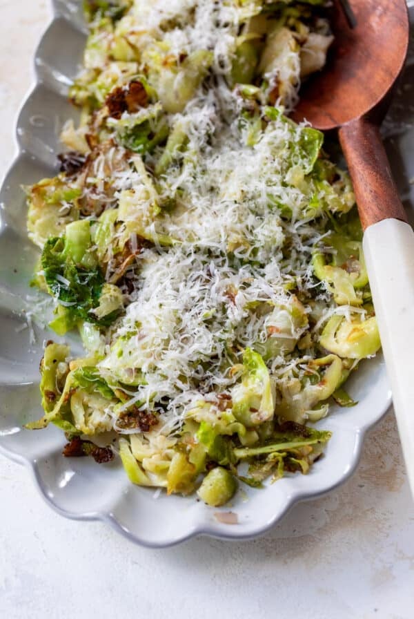 shaved brussels sprouts topped with shredded cheese next to a wooden spoon on a white serving platter.