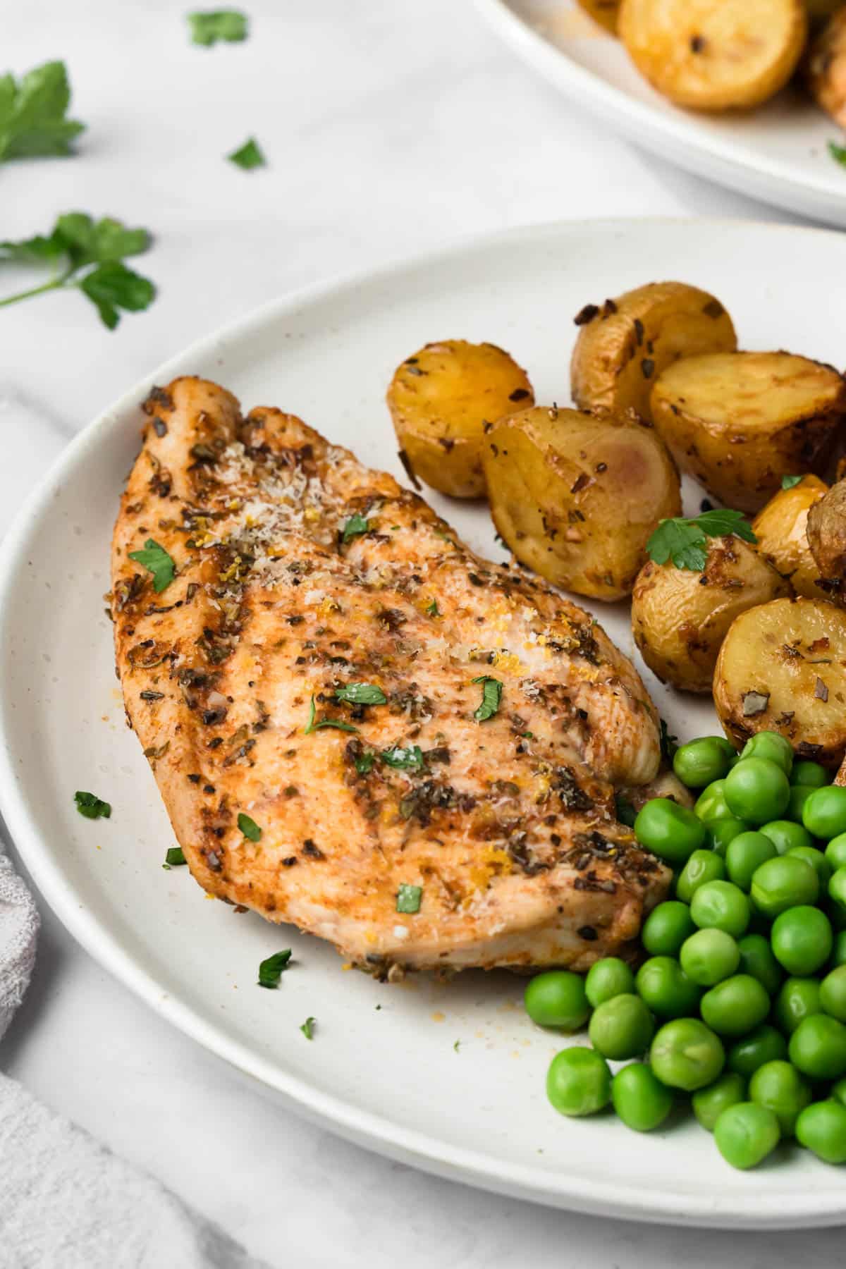 Side view of a roasted chicken breast on a white plate with roasted potatoes and green peas on the side.