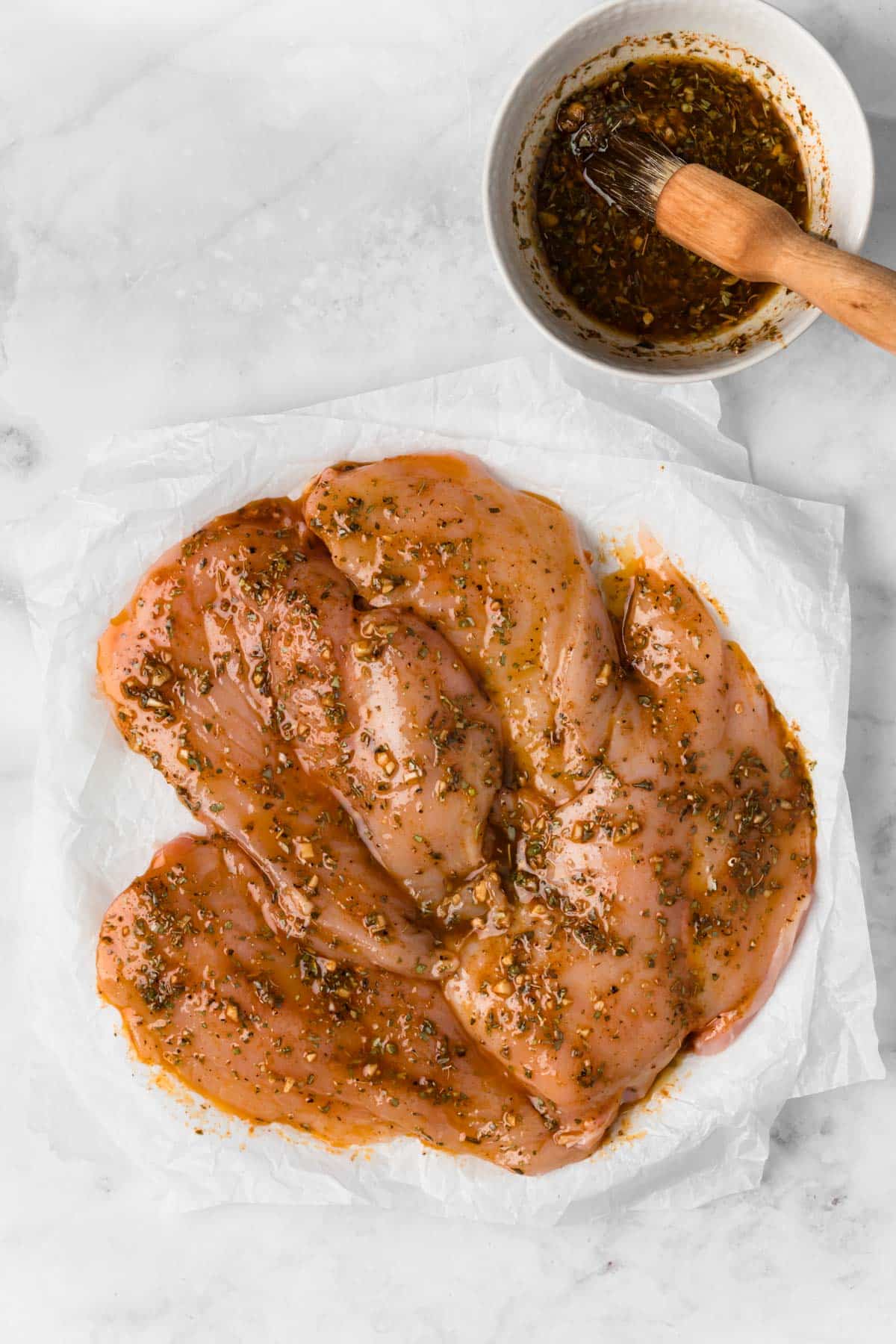 Overhead view of raw chicken breasts on parchment paper next to a small bowl of marinade.