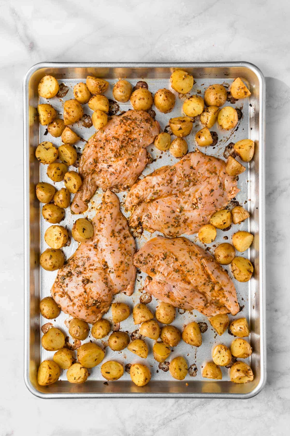 Overhead view of uncooked seasoned chicken breasts and baby potatoes on a sheet pan.