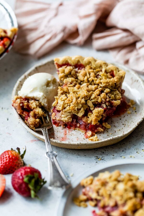 side view of a slice of strawberry crumb pie on a plate with a fork and a scoop of vanilla ice cream