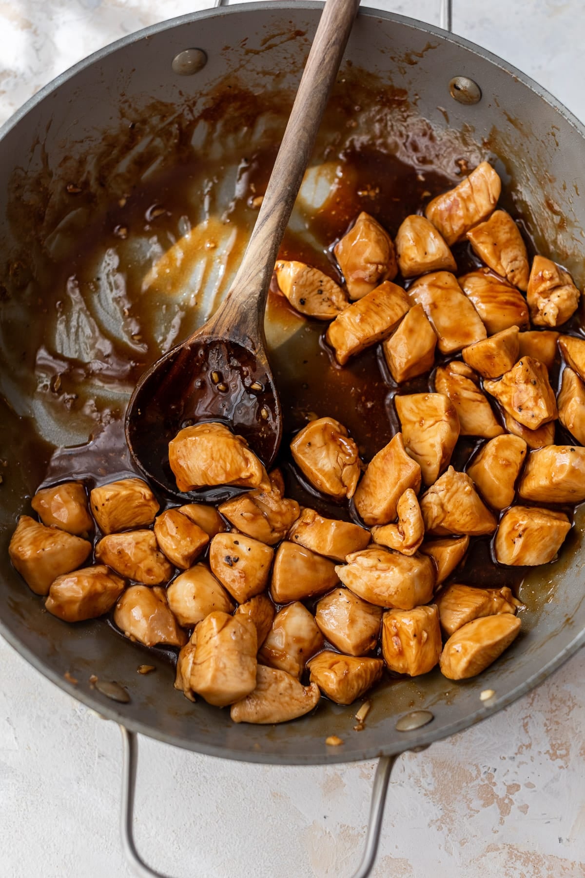 overhead view of a wooden spoon stirring teriyaki sauce-covered cooked chicken bites in a large skillet.