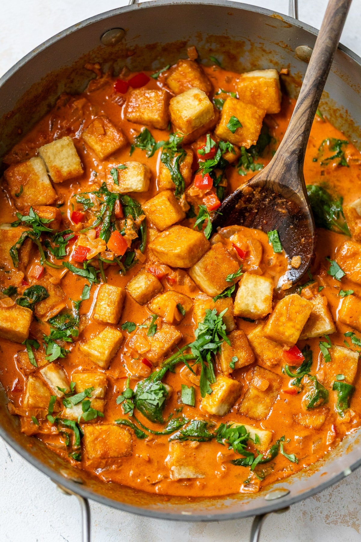 using a wooden spoon to scoop up tofu curry from a large grey skillet.