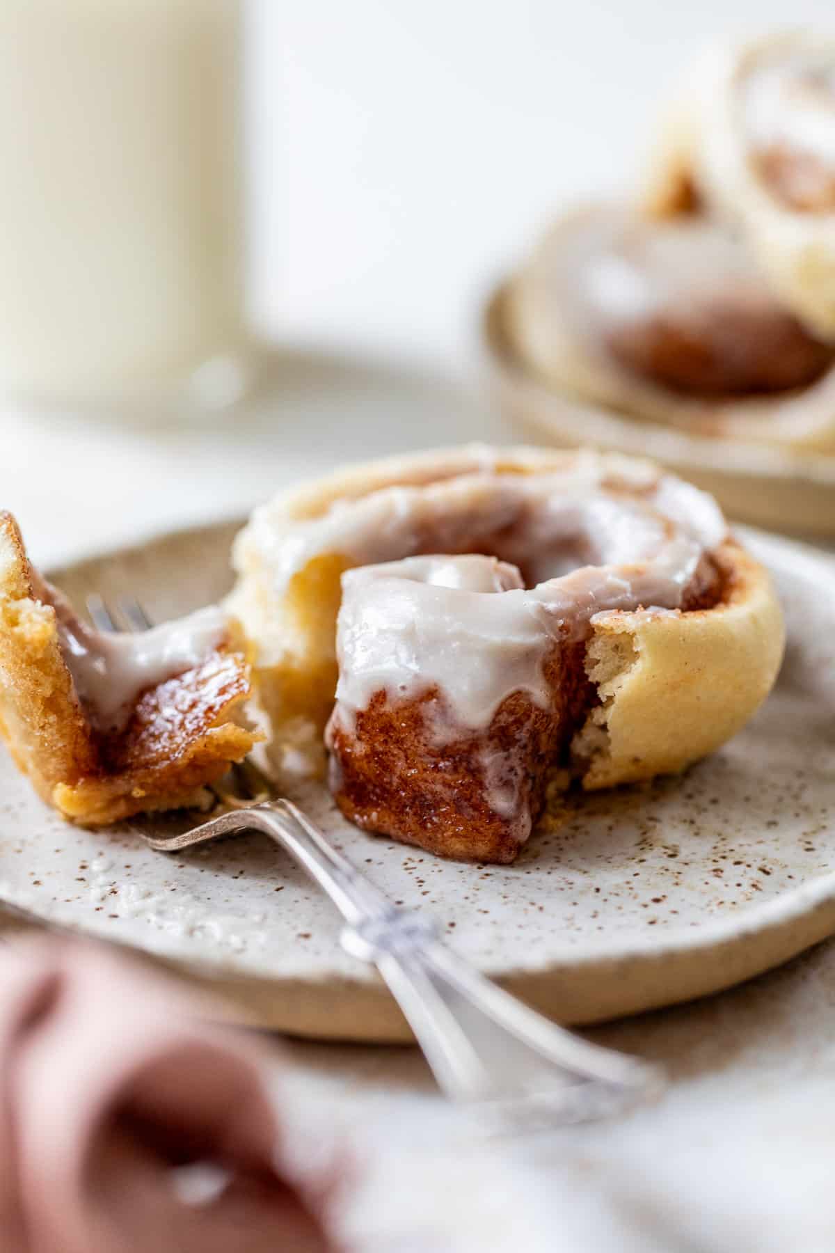 side view of a fork next to a cream cheese frosting-topped vegan cinnamon roll on a plate.