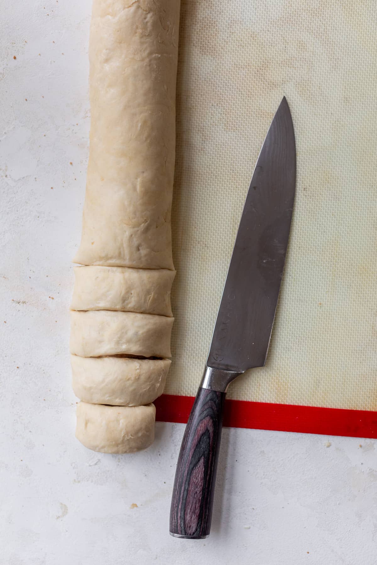 overhead view of a chef's knife next to a log of vegan cinnamon roll dough on a baking mat.