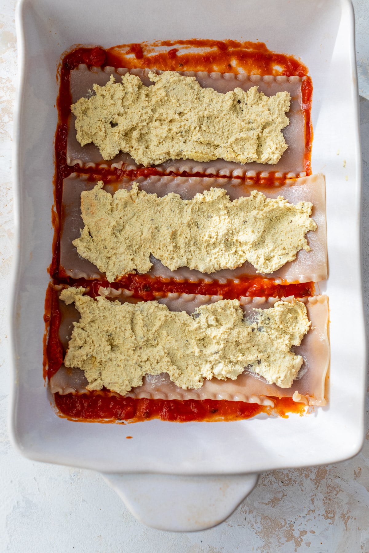 overhead view of lasagna noodles topped with vegan ricotta cheese in a white baking dish.